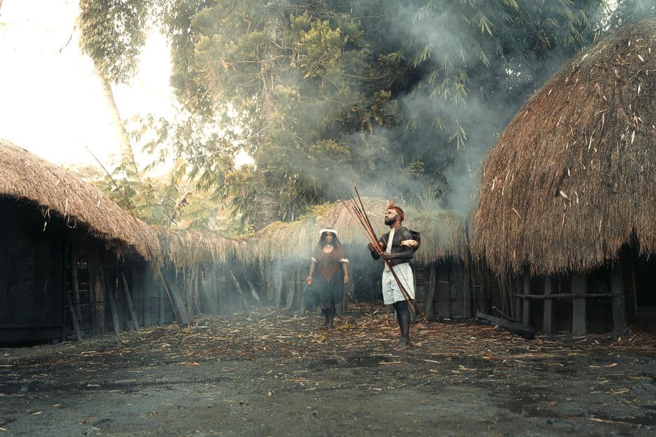Explore a traditional indigenous village with tribal people in ceremonial attire amidst thatched huts and smoke.
