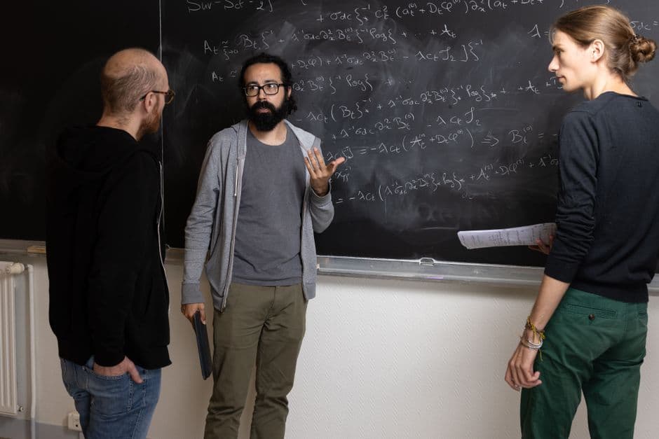 Three adults solving complex mathematical equations on a chalkboard indoors.