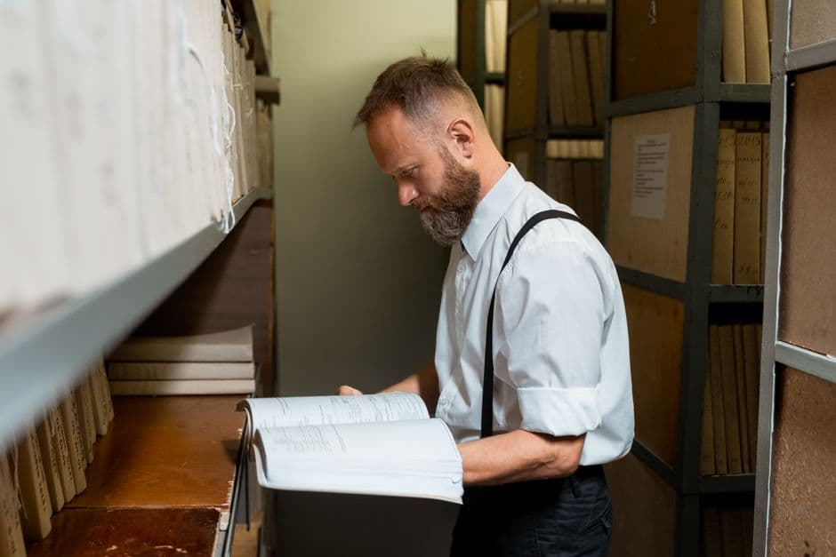 Bearded man in suspenders examining large book in archive room with wooden shelves.