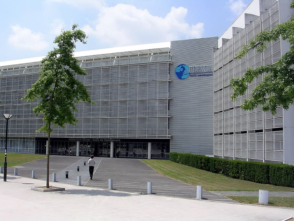 Audencia Business School modern campus facade showing the distinctive glass and metal building with Audencia logo visible against blue sky.