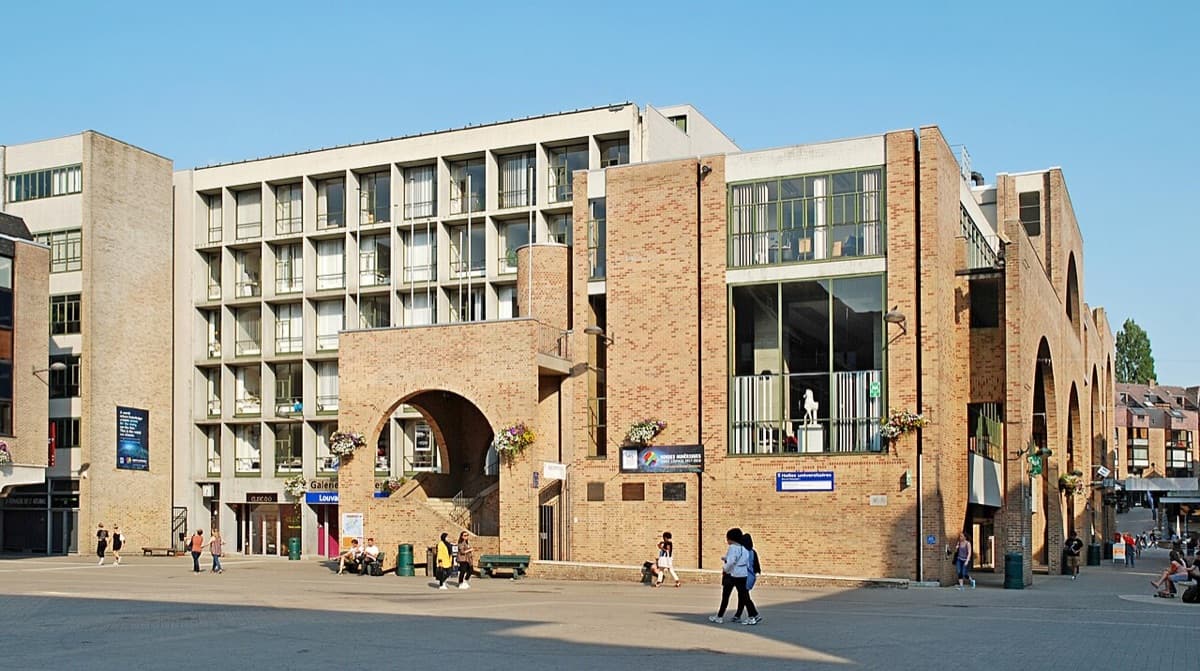 Modern campus building of UCLouvain with brick and white facade, distinctive arched passage, and wide plaza with people on grounds.