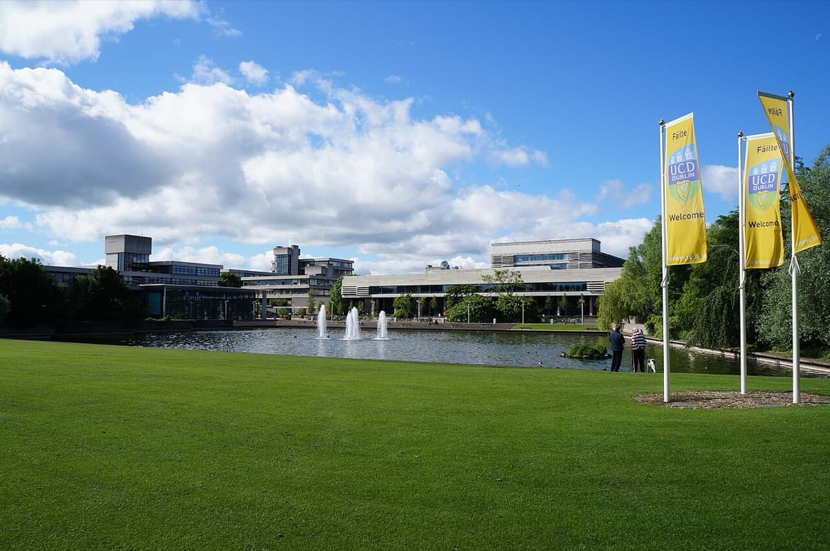 UCD campus showing modern institutional buildings beyond manicured lawns with water feature and yellow welcome banners.