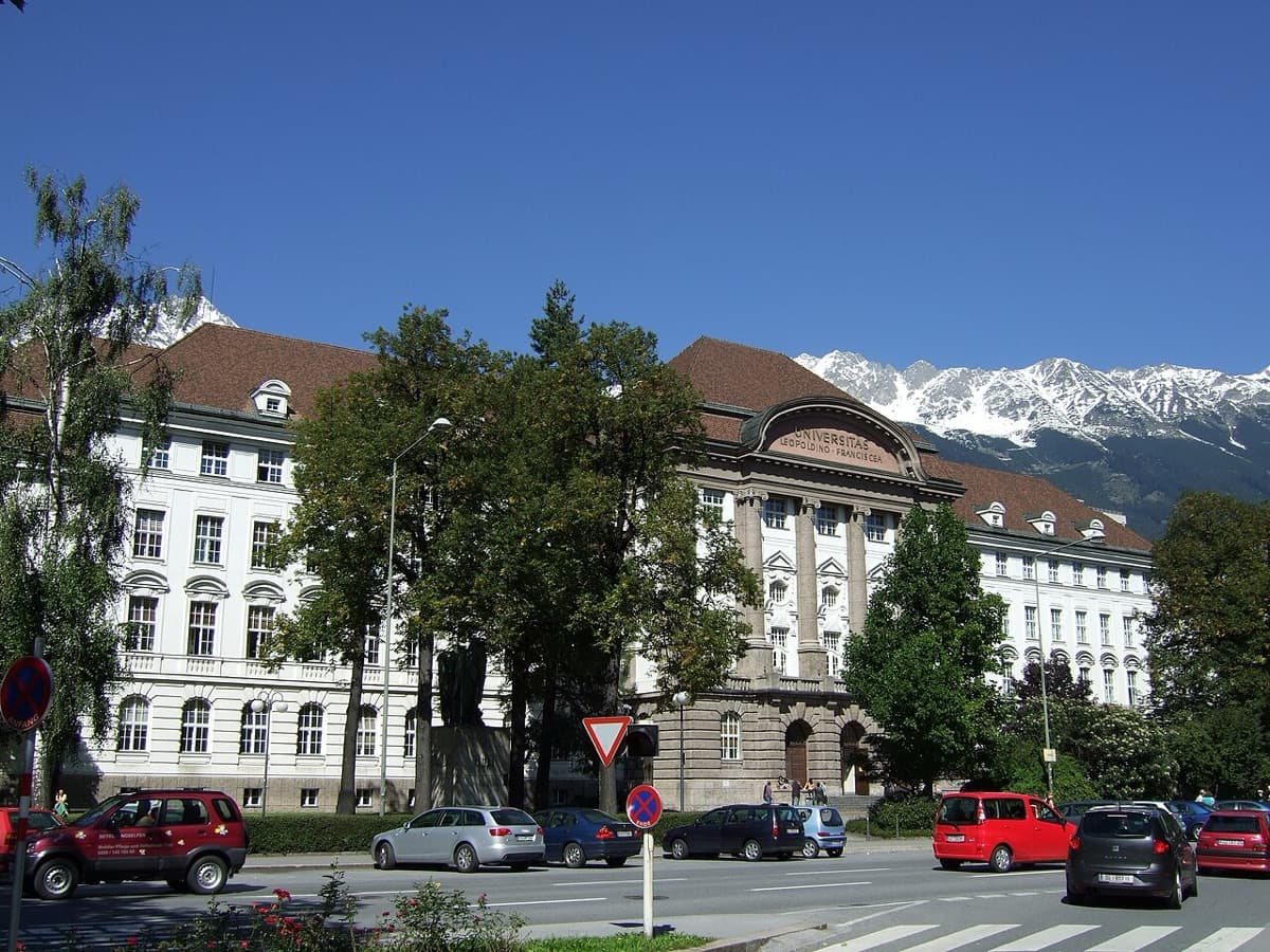 Main building of University of Innsbruck featuring neoclassical white architecture with colonnade and Alpine mountains visible in the background.