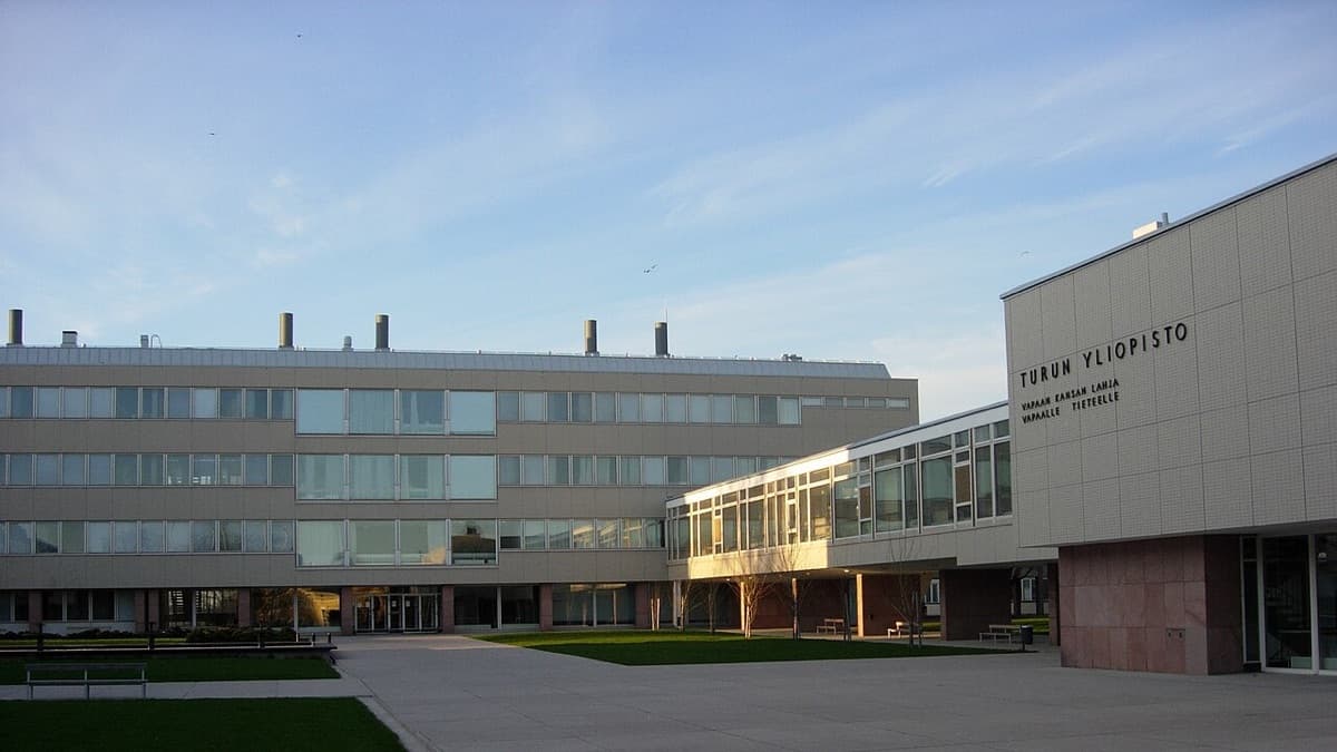 Modern university campus building with clear 'TURUN YLIOPISTO' signage, featuring contemporary architecture with horizontal lines and a central plaza area in daylight.