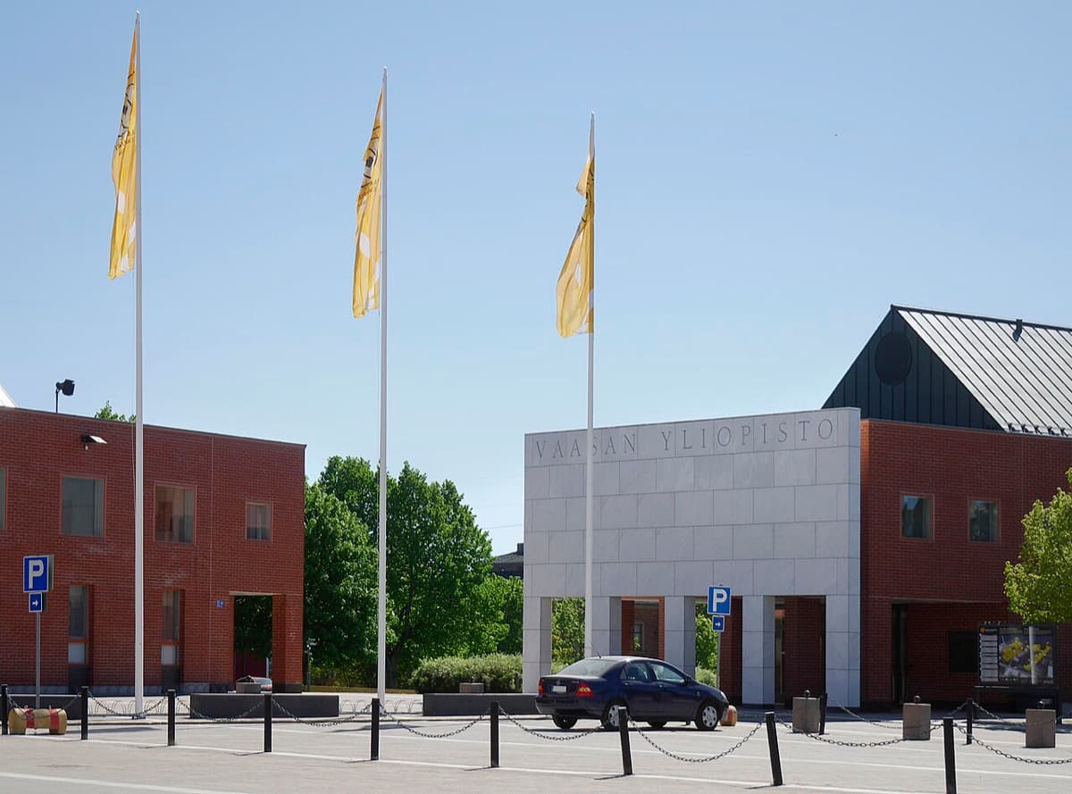 University of Vaasa campus entrance showing modern white and red buildings with 'VAASAN YLIOPISTO' signage and flag poles in daylight.
