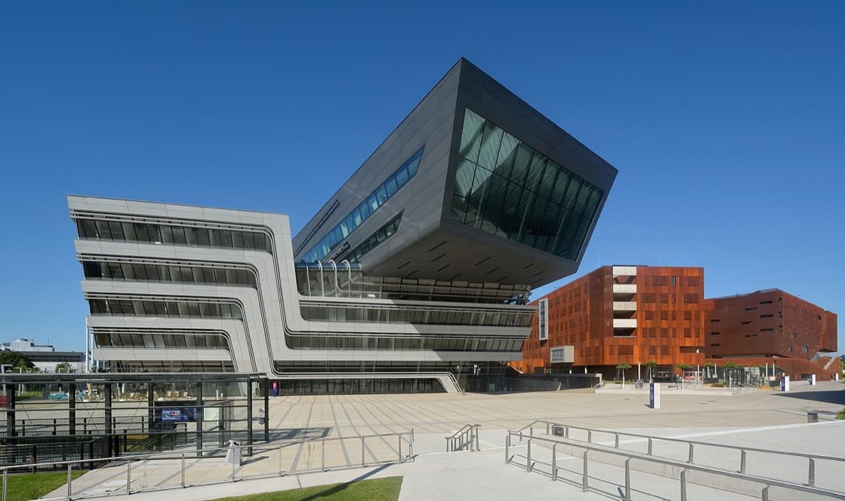Modern dramatic campus architecture of WU Vienna featuring a distinctive cantilevered section with white and dark facade elements and wide plaza.